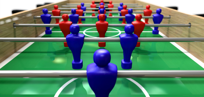 A perspective view of a wooden foosball table showing a blue and red team on a green marked pitch on an isolated white background
