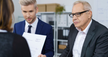 Three serious mature and young workers discussing business in small office with bookshelf in background