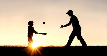 silhouette of a father and his young child playing baseball outside, isolated against the sunsetting sky on a summer day.