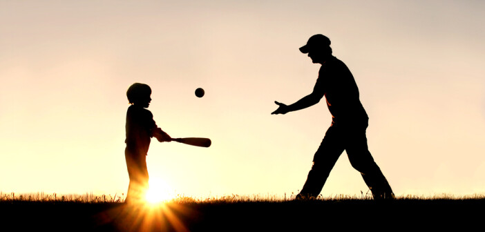 silhouette of a father and his young child playing baseball outside, isolated against the sunsetting sky on a summer day.